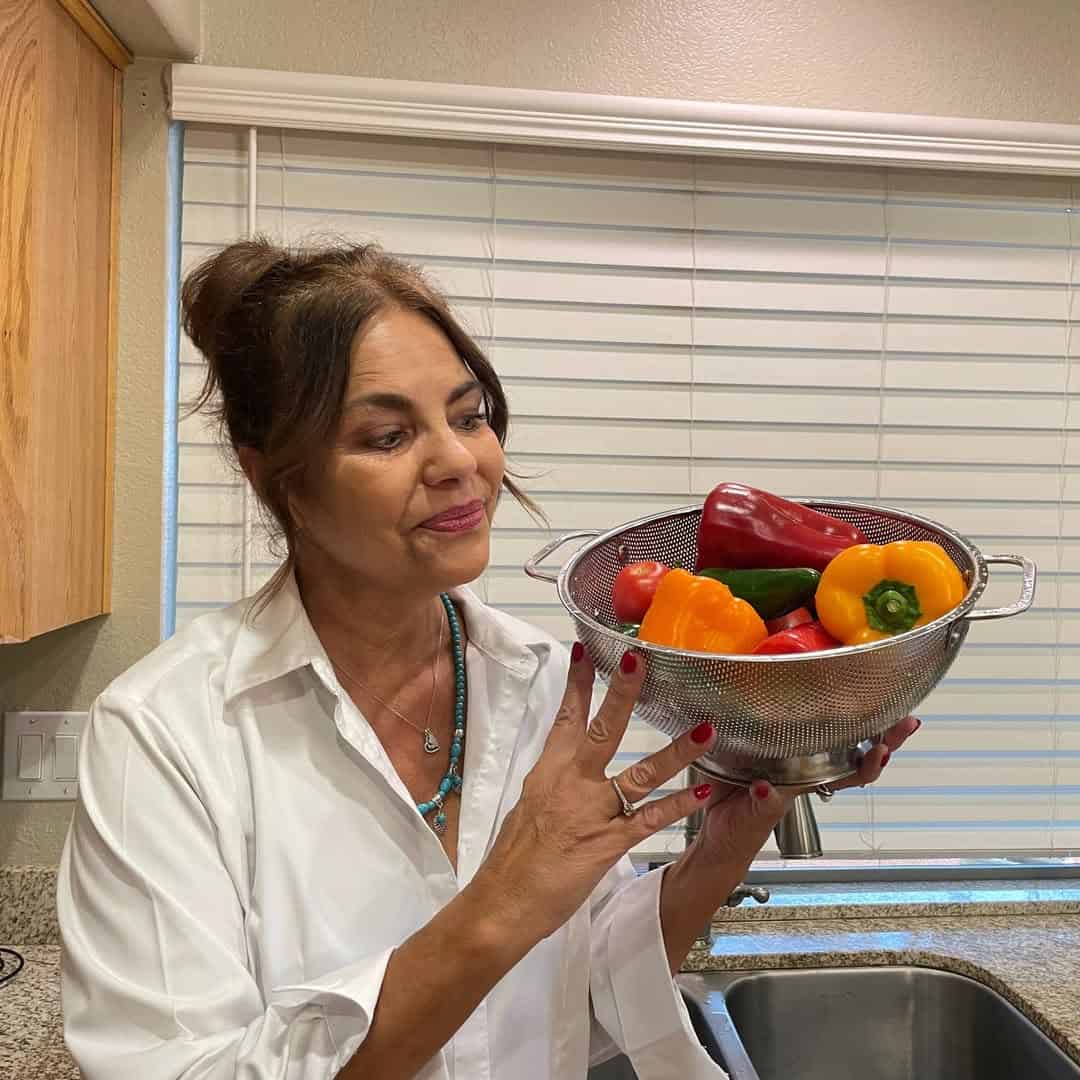 A woman in a kitchen holds a metal colander—one of the essential cooking tools for beginners—filled with colorful bell peppers and tomatoes.