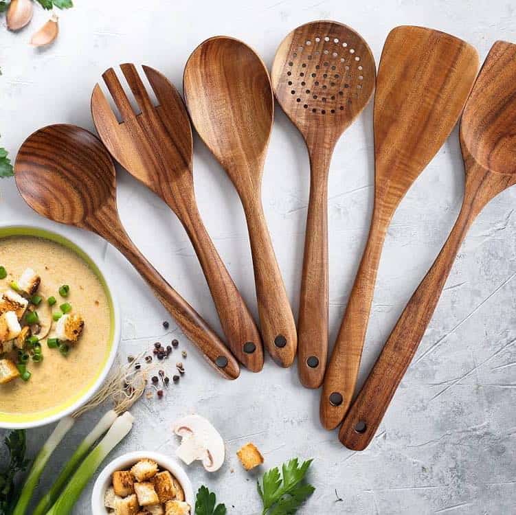 Six essential cooking tools for beginners—wooden kitchen utensils—are arranged on a gray surface near a bowl of soup with croutons and vegetables.