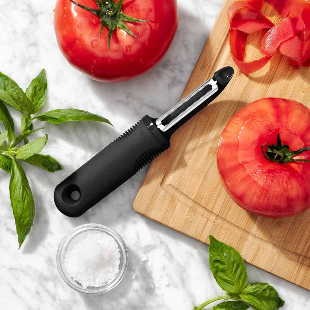 A black vegetable peeler, essential cooking tools for beginners, sits among tomatoes, basil, and salt on a marble countertop with a wooden cutting board.