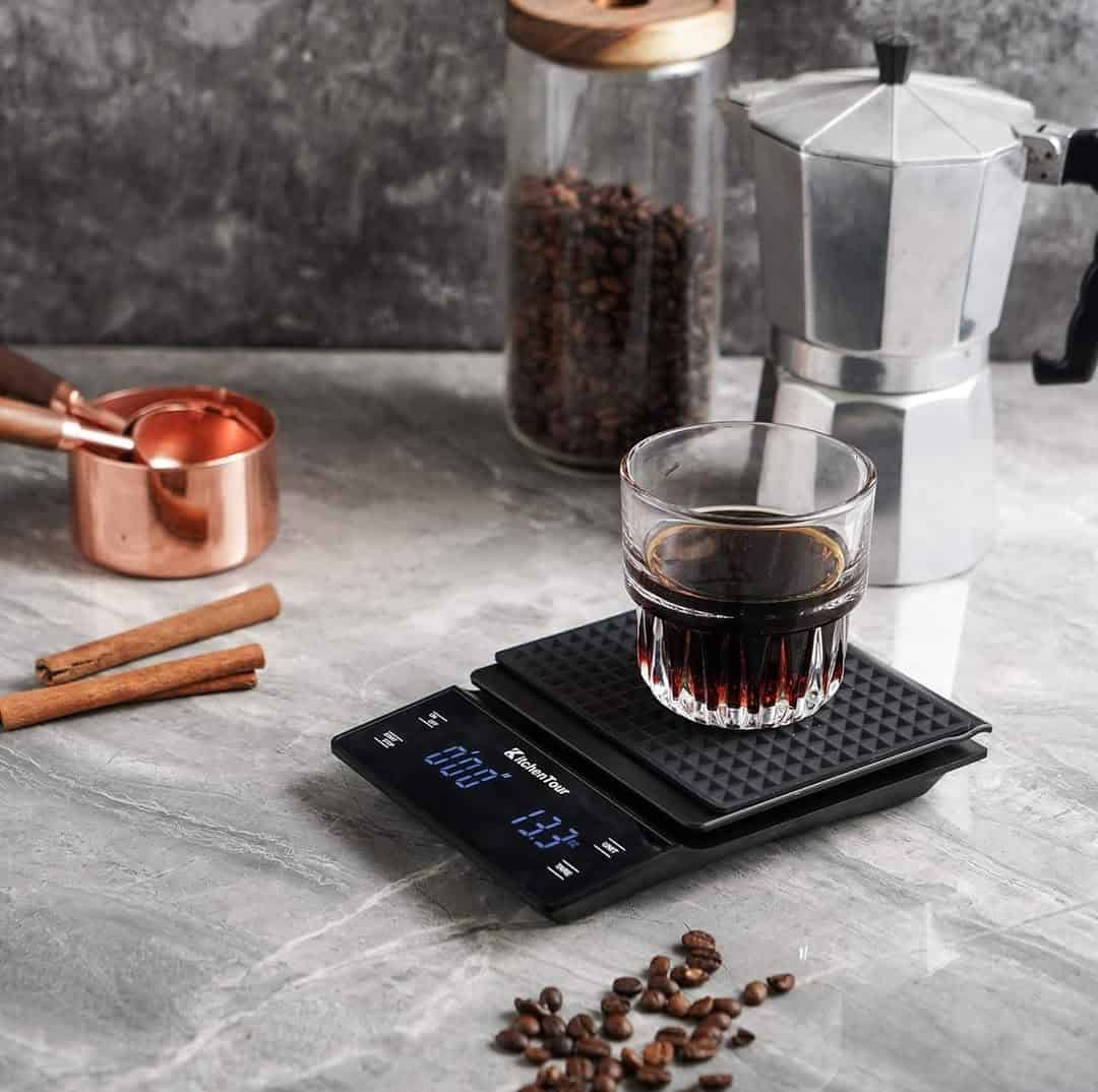 A glass of coffee on a digital scale, with coffee beans, a moka pot, and cinnamon sticks nearby.