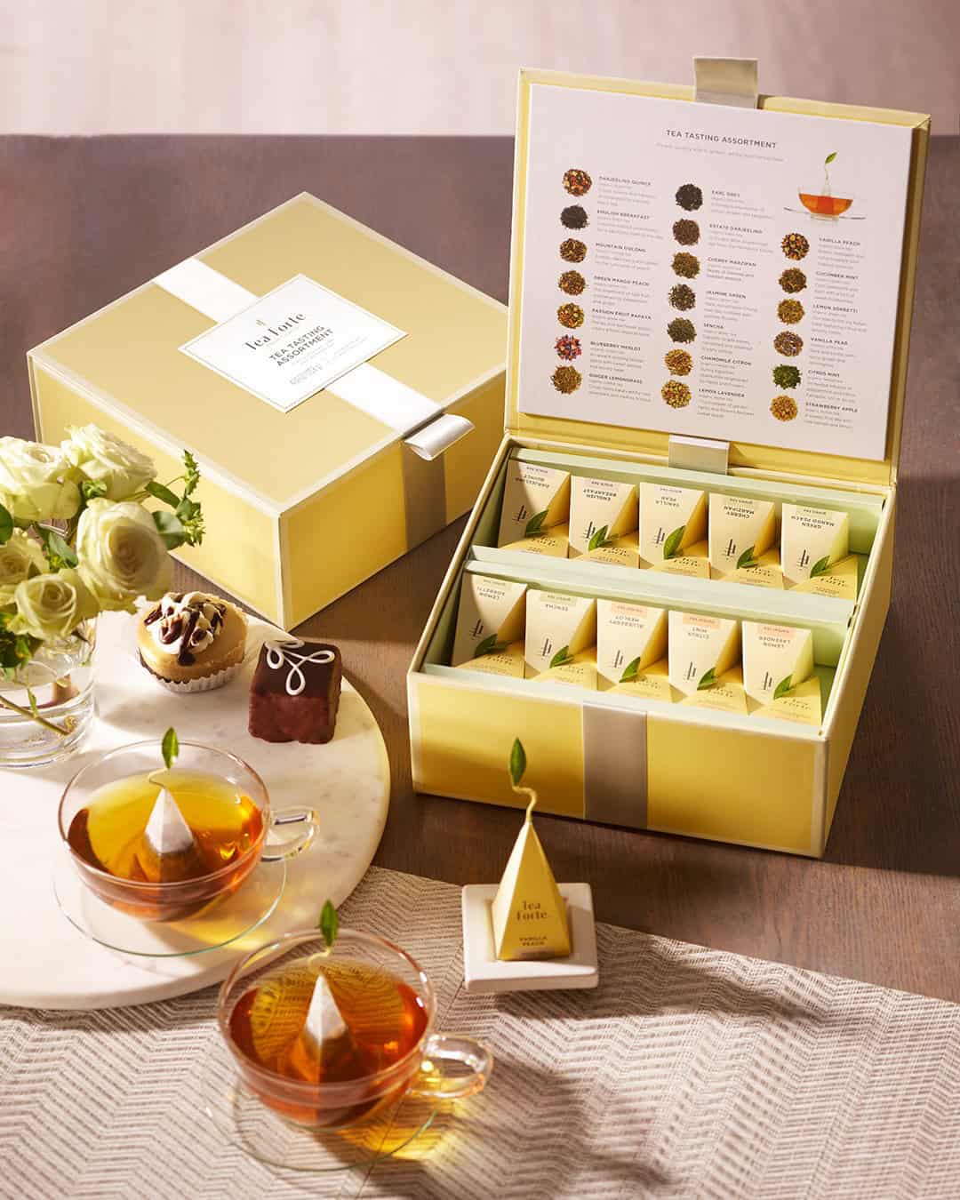 Elegant yellow tea gift box with pyramid tea bags, two cups of tea, and assorted desserts on a wooden table.
