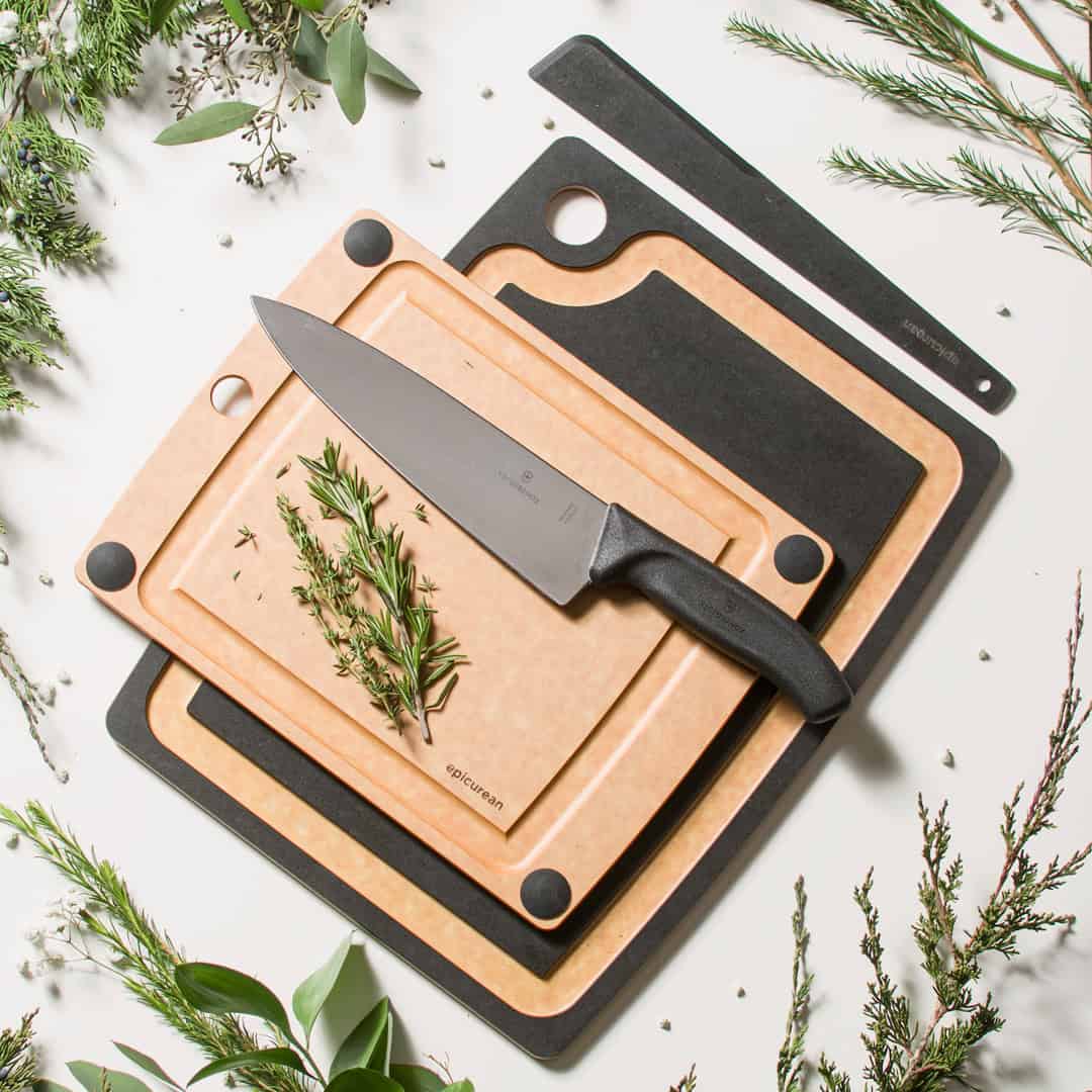 Three cutting boards stacked with a chef’s knife, herbs, and greenery arranged around them on a white background—essential cooking tools for beginners.