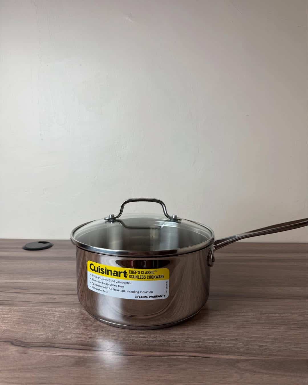 A stainless steel Cuisinart saucepan with lid, one of the essential cooking tools for beginners, sits on a wooden surface against a plain wall.