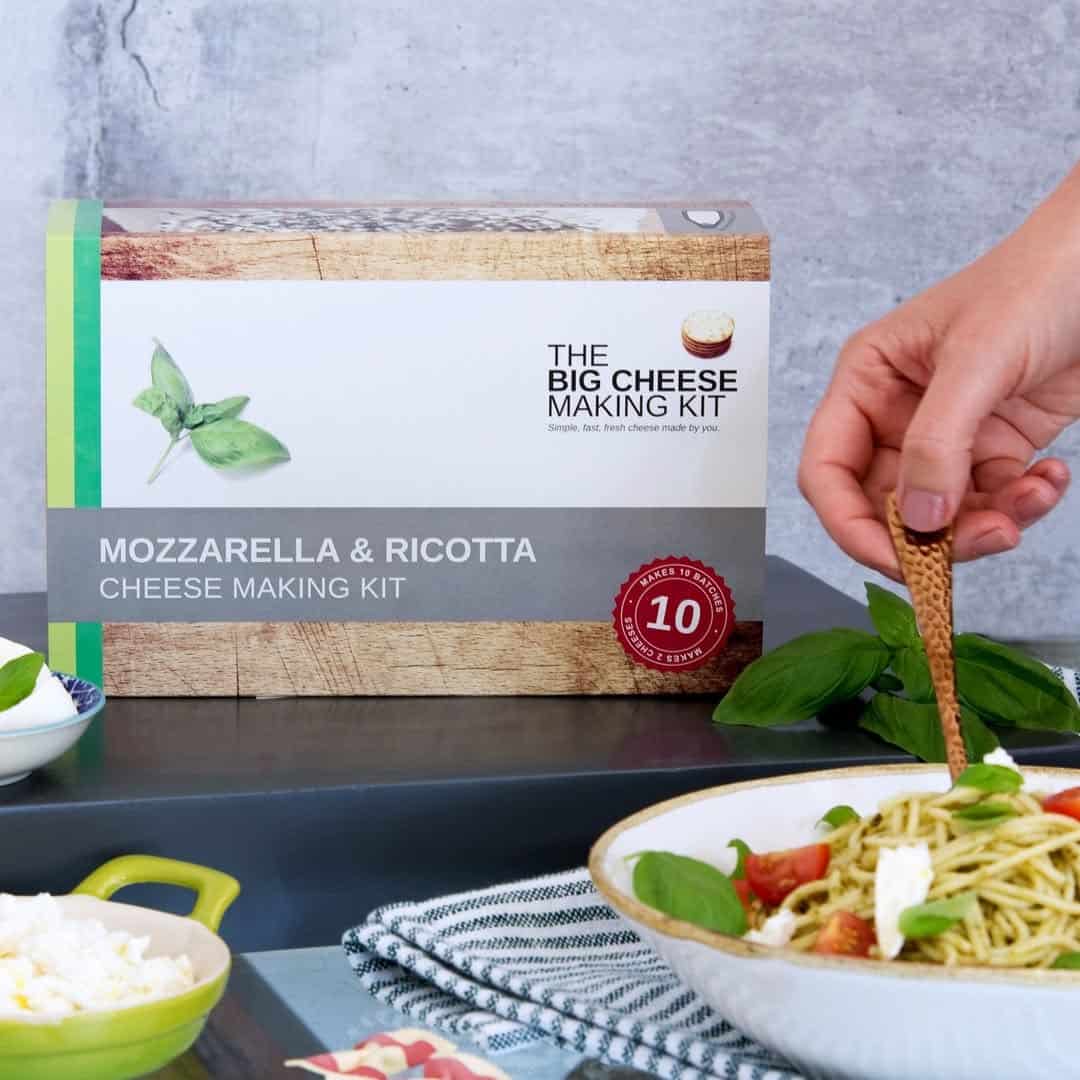 A hand sprinkles cheese on pasta in front of a Mozzarella & Ricotta Cheese Making Kit box—perfect for gifts for cheese lovers.