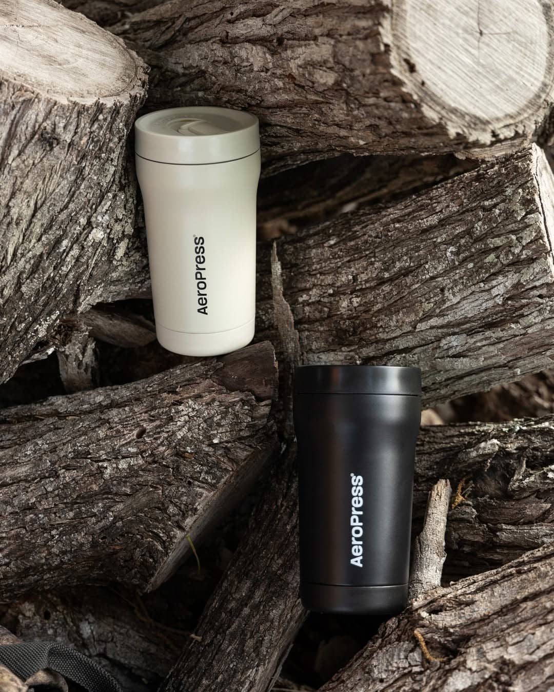 Two AeroPress travel mugs, one white and one black, placed on stacked firewood outdoors.