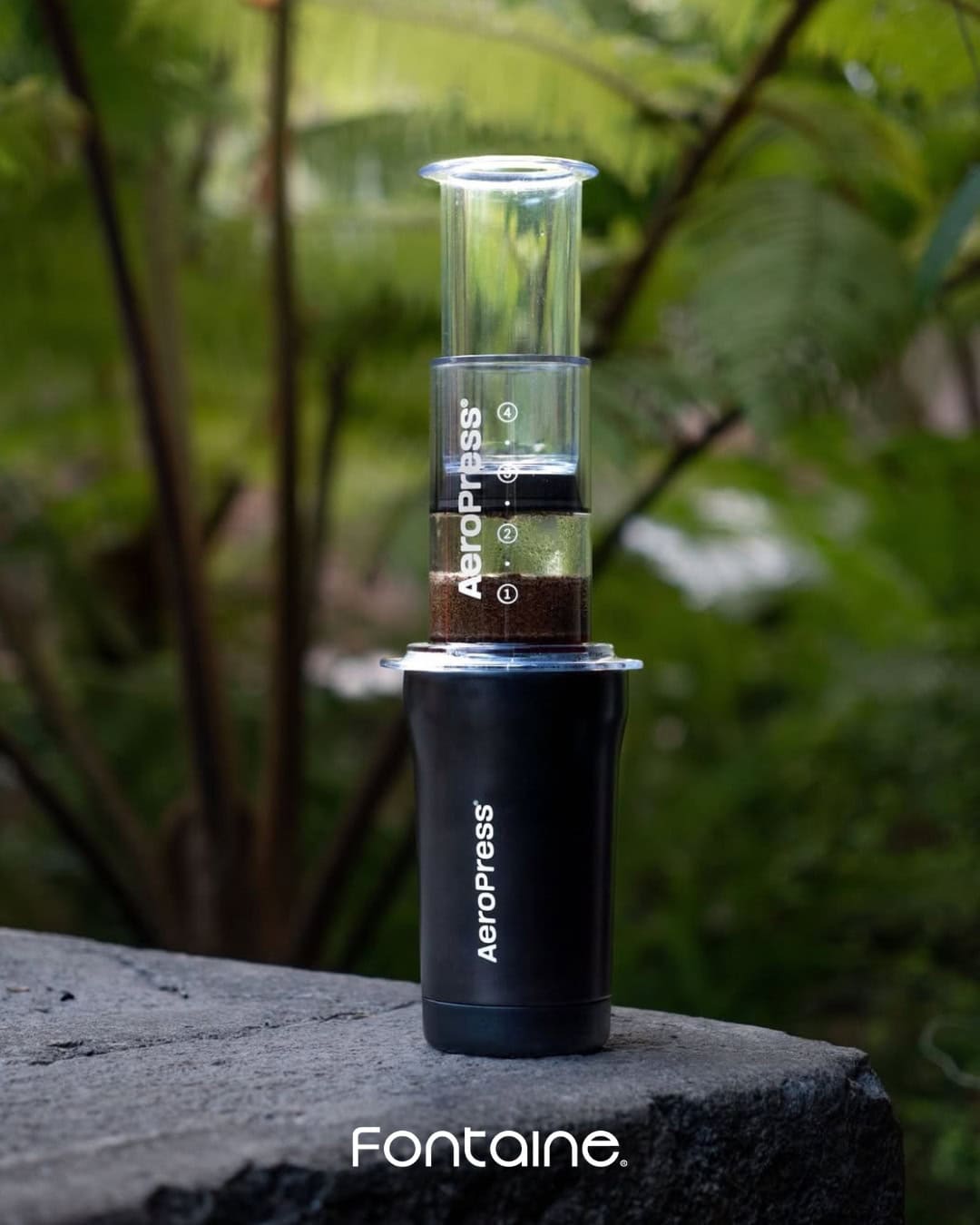 An AeroPress coffee maker on a stone surface outdoors, with greenery in the background.