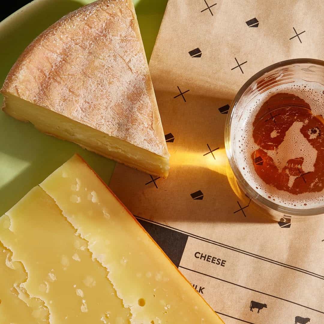 A wedge of cheese, a glass of beer, and a paper wrapper on a green plate in sunlight-perfect gifts for cheese lovers to enjoy a relaxing afternoon.