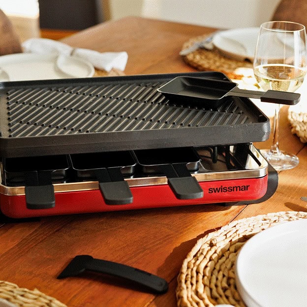 A red Swissmar raclette grill sits on a wooden table set for a meal, with plates and a glass of wine—perfect for gifts for cheese lovers.