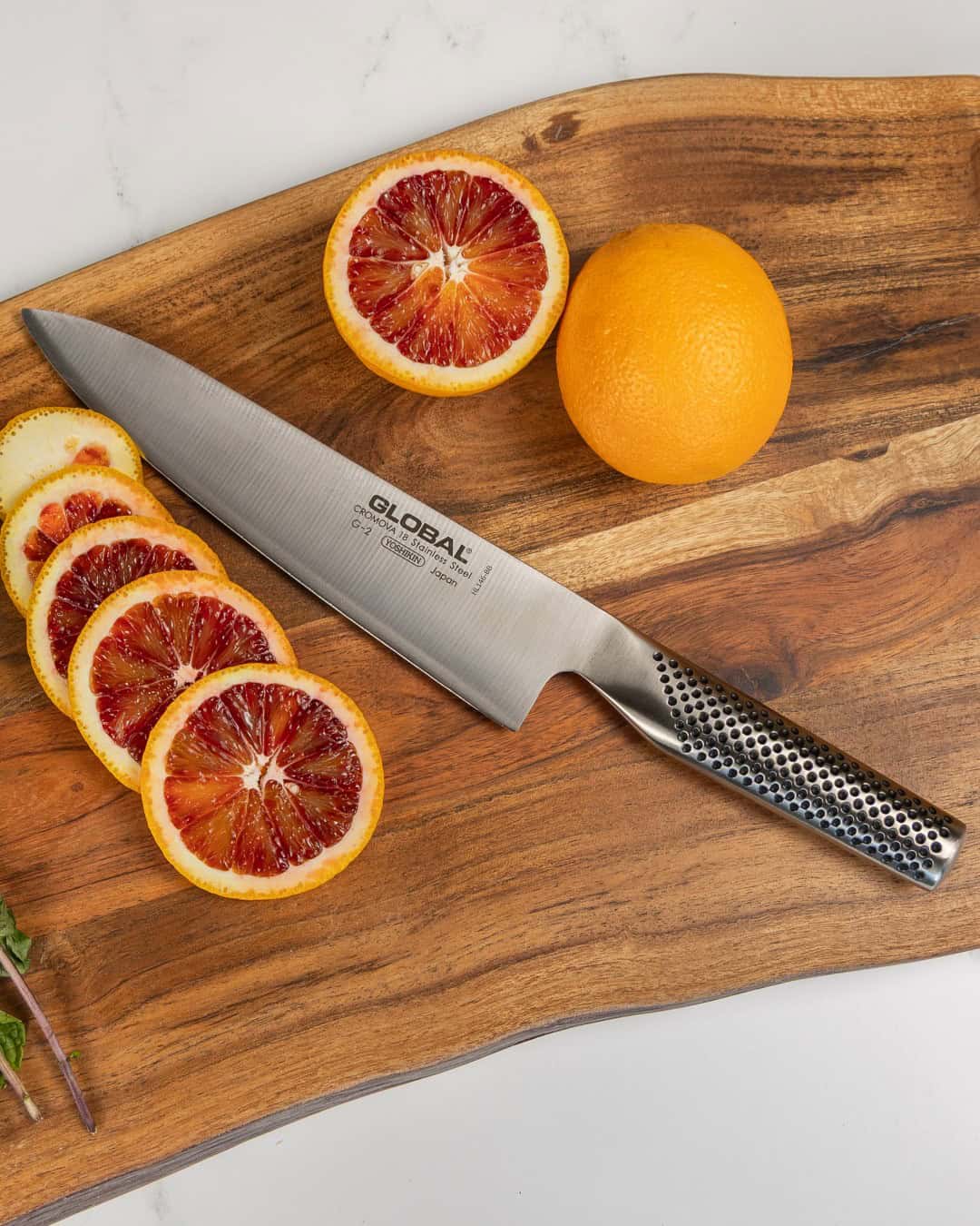 A chef's knife-one of the essential cooking tools for beginners-rests on a wooden board beside sliced and whole blood oranges.