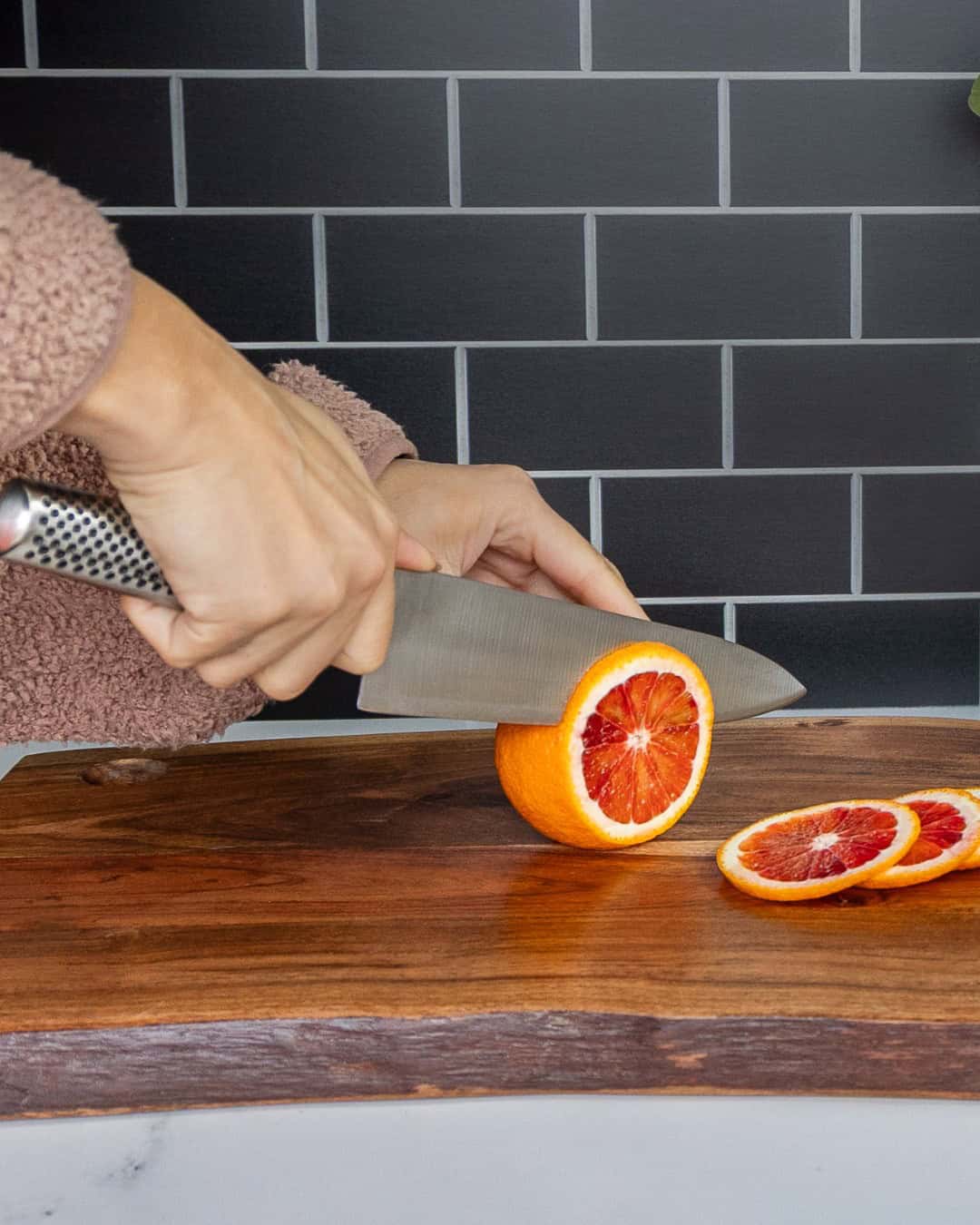 A person slices a blood orange with a knife, one of the essential cooking tools for beginners, on a wooden cutting board in a kitchen.