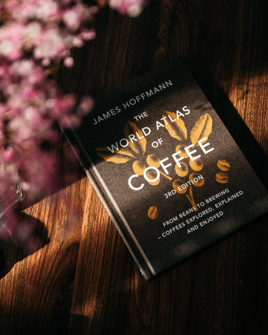 A book titled "The World Atlas of Coffee" by James Hoffmann rests on a wooden table with pink flowers nearby.
