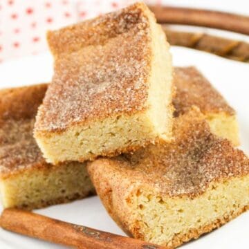 Three cinnamon sugar blondies stacked on a white plate, with a cinnamon stick in front.