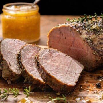 Sliced roasted pork loin with herbs on a wooden board, with a jar of mustard in the background.