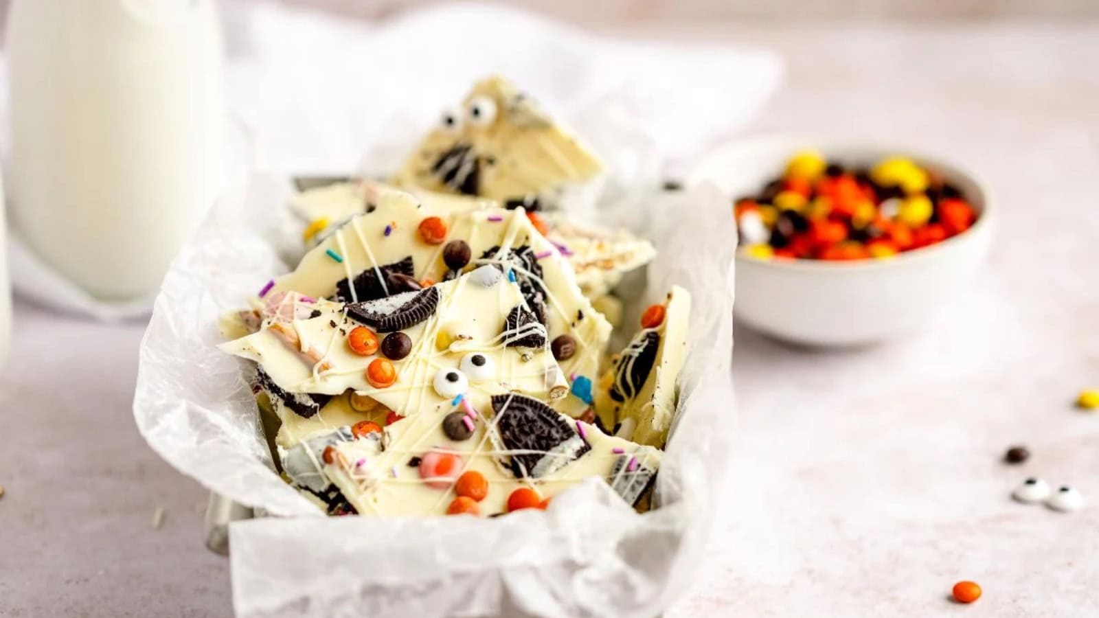 White chocolate Halloween candy bark topped with cookie pieces, candy eyes, and colorful sprinkles in a paper-lined dish.