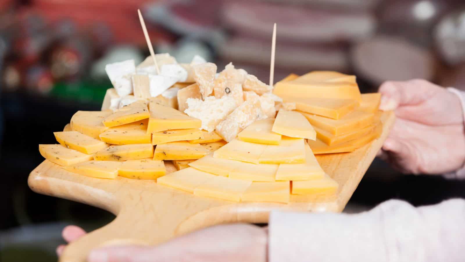 A person holds a wooden board with assorted sliced and cubed cheeses on display—perfect gifts for cheese lovers.