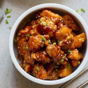 A bowl of glazed chicken pieces topped with sesame seeds and chopped chives, with chopsticks beside the bowl.