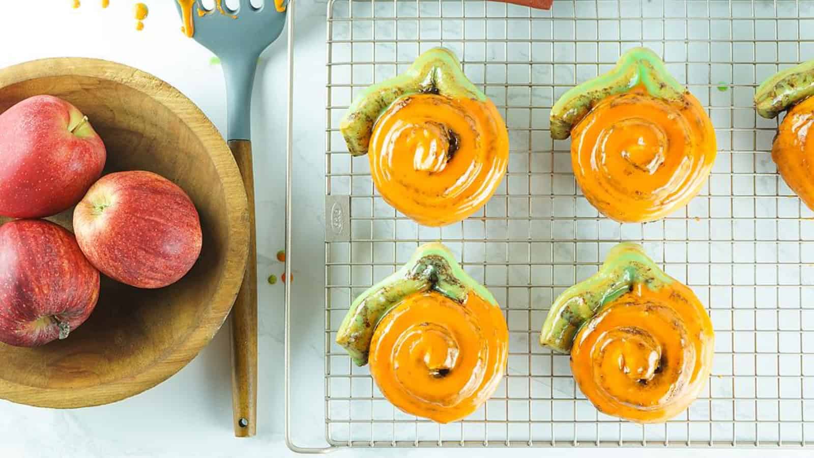 Pumpkin-shaped cinnamon rolls with orange icing cooling on a wire rack next to a bowl of apples.