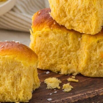 Three fluffy, golden Hawaiian rolls stacked on a wooden board, topped with flaky salt.