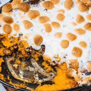 Baked sweet potato casserole topped with toasted marshmallows in a round dish, partially eaten, with a metal serving spoon visible.
