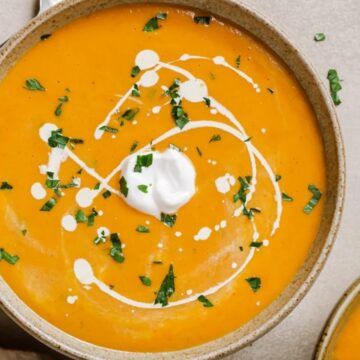 Bowl of orange soup with cream swirls and chopped herbs on top, viewed from above.