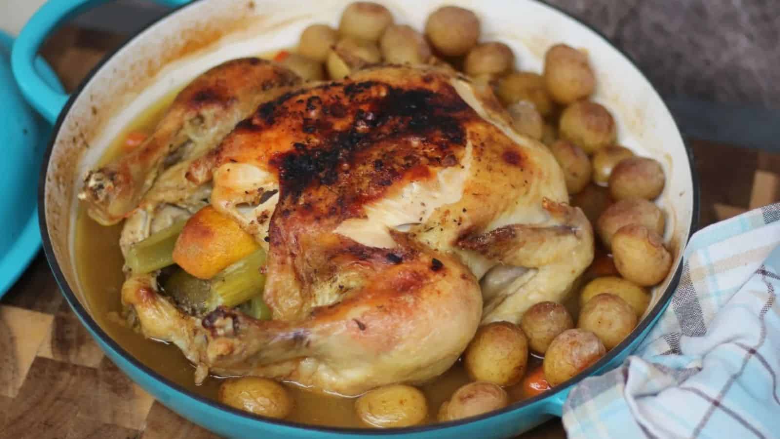 Roast chicken with crispy skin, surrounded by baby potatoes and vegetables in a blue baking dish.