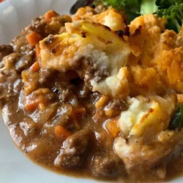 A serving of shepherd’s pie with ground meat, vegetables, and mashed potatoes on a white plate with salad.