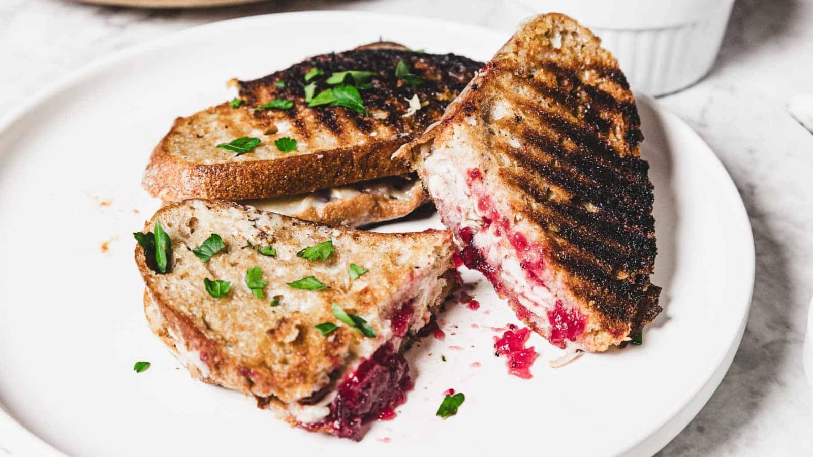 Close-up of a grilled sandwich with turkey, cranberry sauce, and herbs on toasted bread.