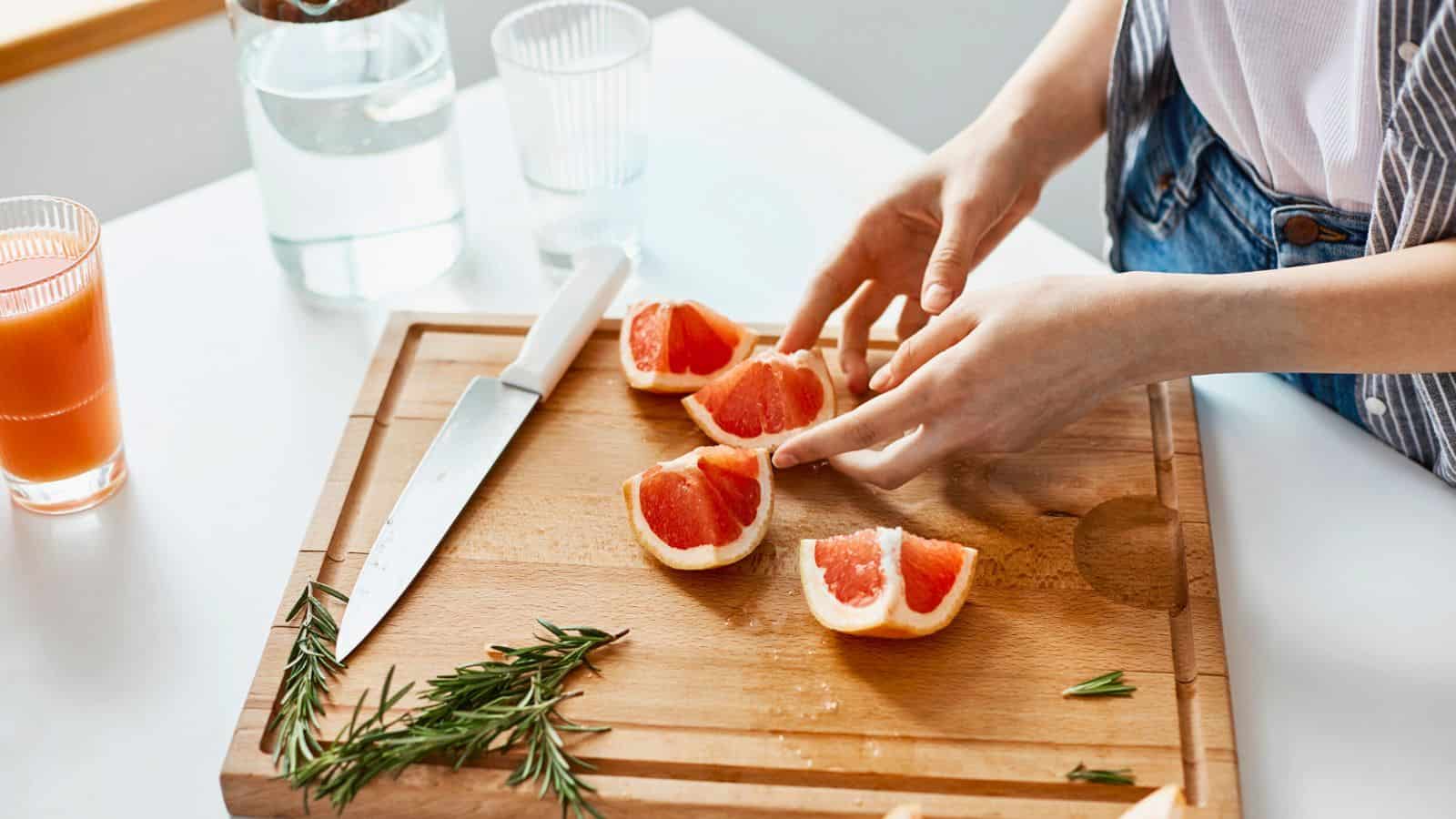 Person slicing grapefruit on a wooden cutting board with a knife, rosemary, and a glass of juice nearby—perfect inspiration for thoughtful gifts for cheese lovers who appreciate fresh and flavorful pairings.