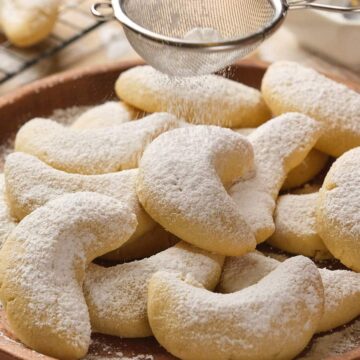 A plate of crescent-shaped cookies being dusted with powdered sugar using a small sieve.