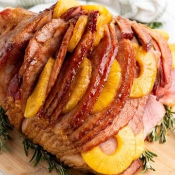 Sliced glazed ham garnished with pineapple rings and rosemary on a wooden cutting board.