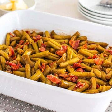 A white baking dish filled with cooked green beans and pieces of bacon, sitting on a cooling rack.