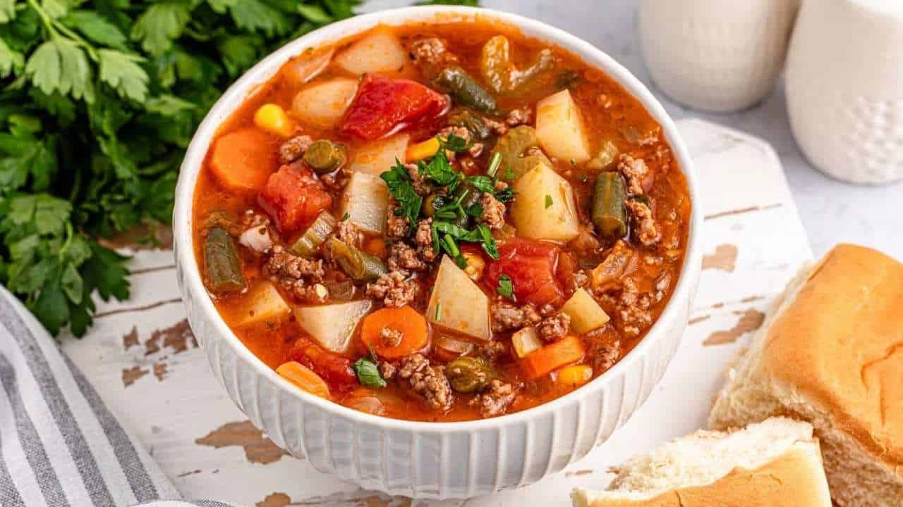 A bowl of hearty vegetable soup with ground meat, potatoes, carrots, corn, green beans, and tomatoes. It is garnished with fresh herbs. The bowl is on a white surface with two bread rolls and a bunch of parsley nearby.