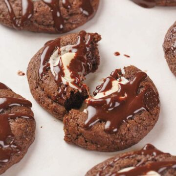 Chocolate cookies with cream centers, drizzled with chocolate, on a white surface; one cookie is broken in half.