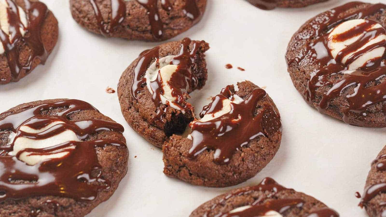 Chocolate cookies with cream centers, drizzled with chocolate, on a white surface; one cookie is broken in half.