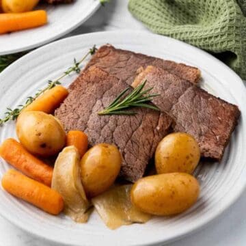Plate with sliced pot roast, baby potatoes, carrots, onions, and a sprig of rosemary for garnish.