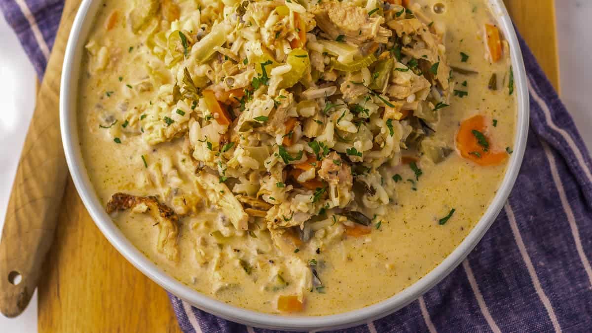 Bowl of creamy turkey and wild rice soup with herbs on a wooden board and a striped cloth napkin.