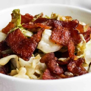 Bowl of cooked cabbage topped with crispy bacon pieces on a white surface.