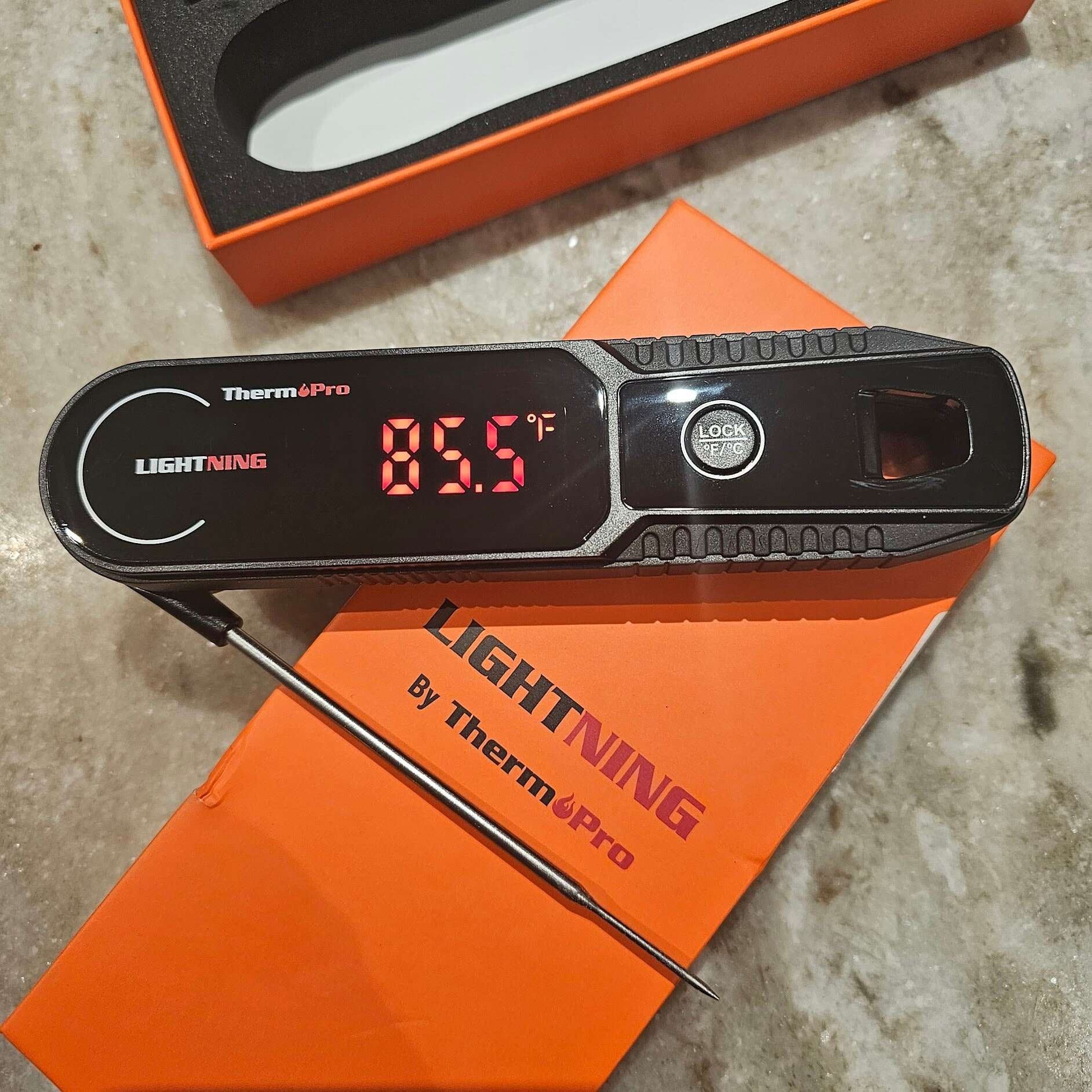 A digital meat thermometer, one of the essential cooking tools for beginners, displays 85.5°F as it rests on an orange ThermoPro box.