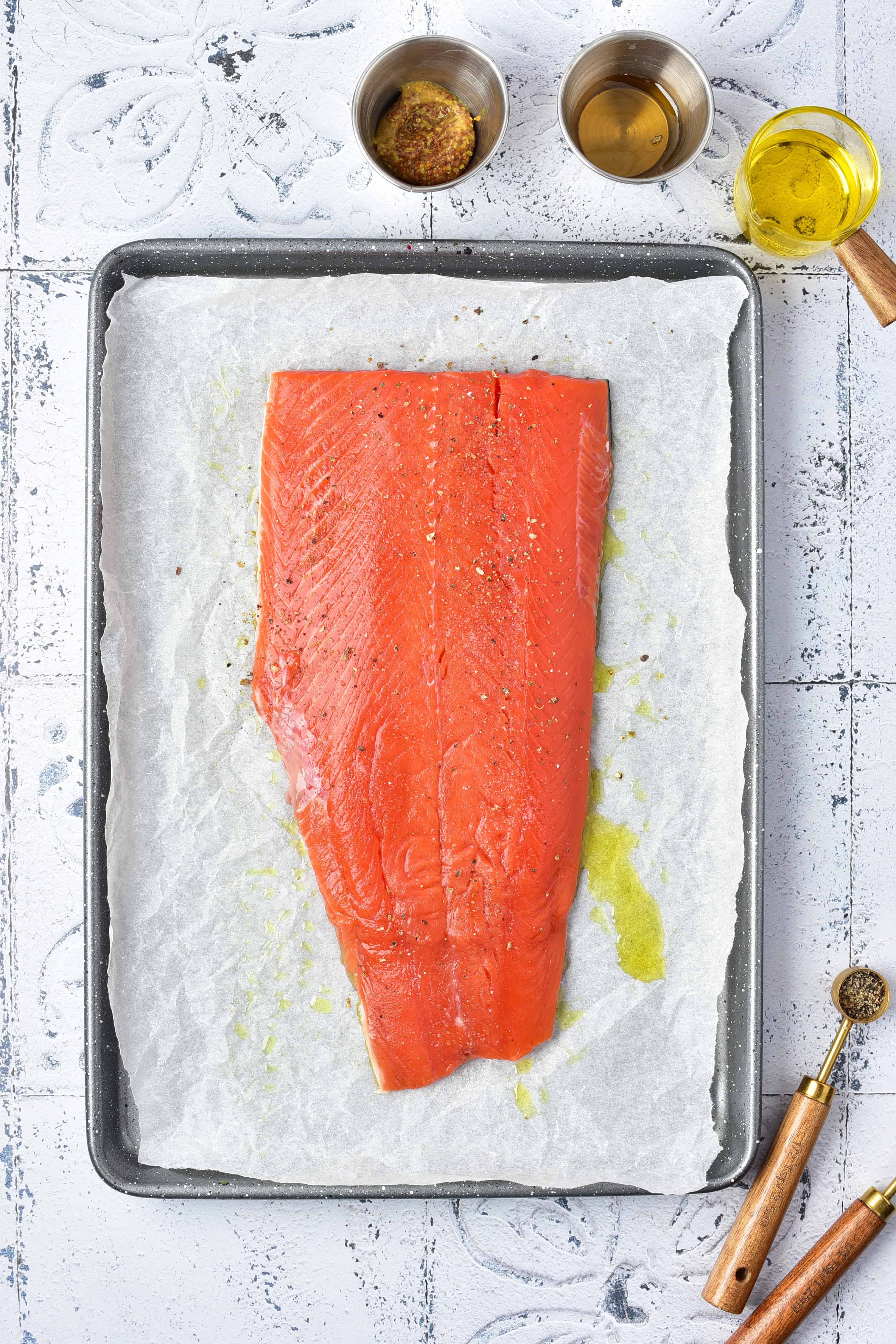 Raw salmon fillet on parchment-lined tray, with oil, mustard, and seasoning in small bowls nearby.