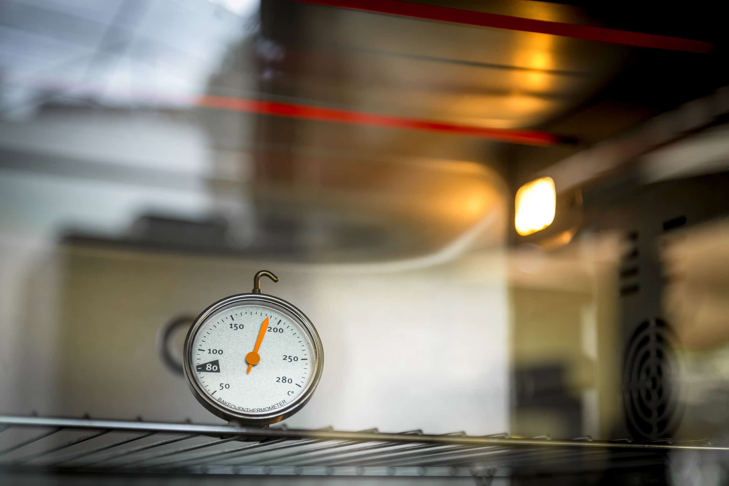 A metal oven thermometer, one of the essential kitchen gadgets for holiday cooking, sits on a wire rack inside a lit oven, displaying a temperature reading.