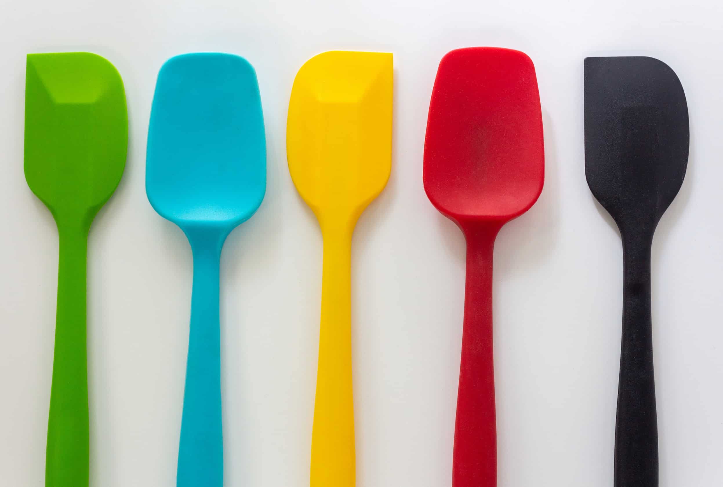 Five silicone spatulas in green, blue, yellow, red, and black arranged in a row on a white background&mdash;essential kitchen tools for holiday baking.