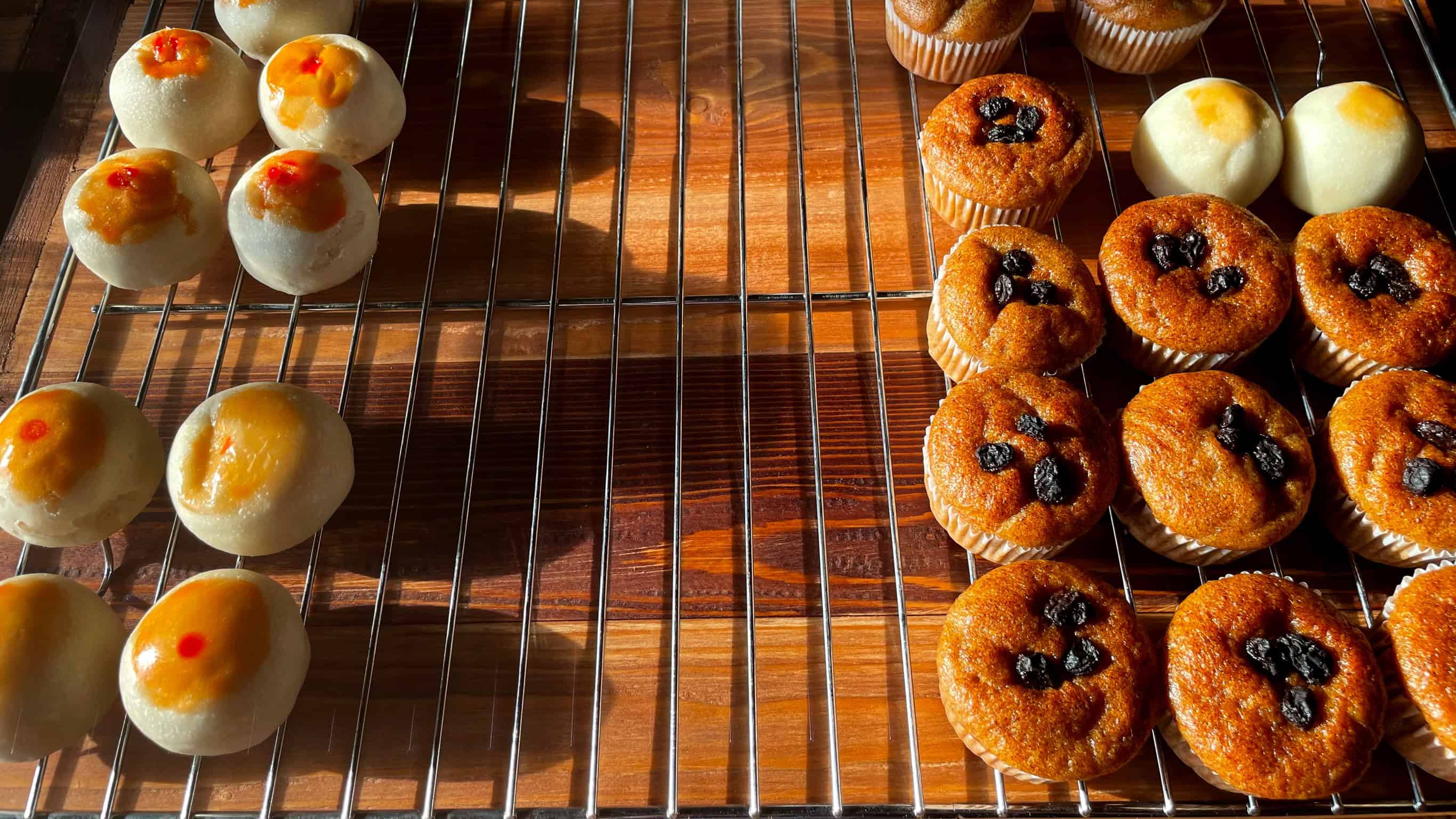 Pastries and muffins cooling on a wire rack, with sunlight casting shadows on the wooden surface&mdash;perfect inspiration for choosing kitchen tools for holiday baking.