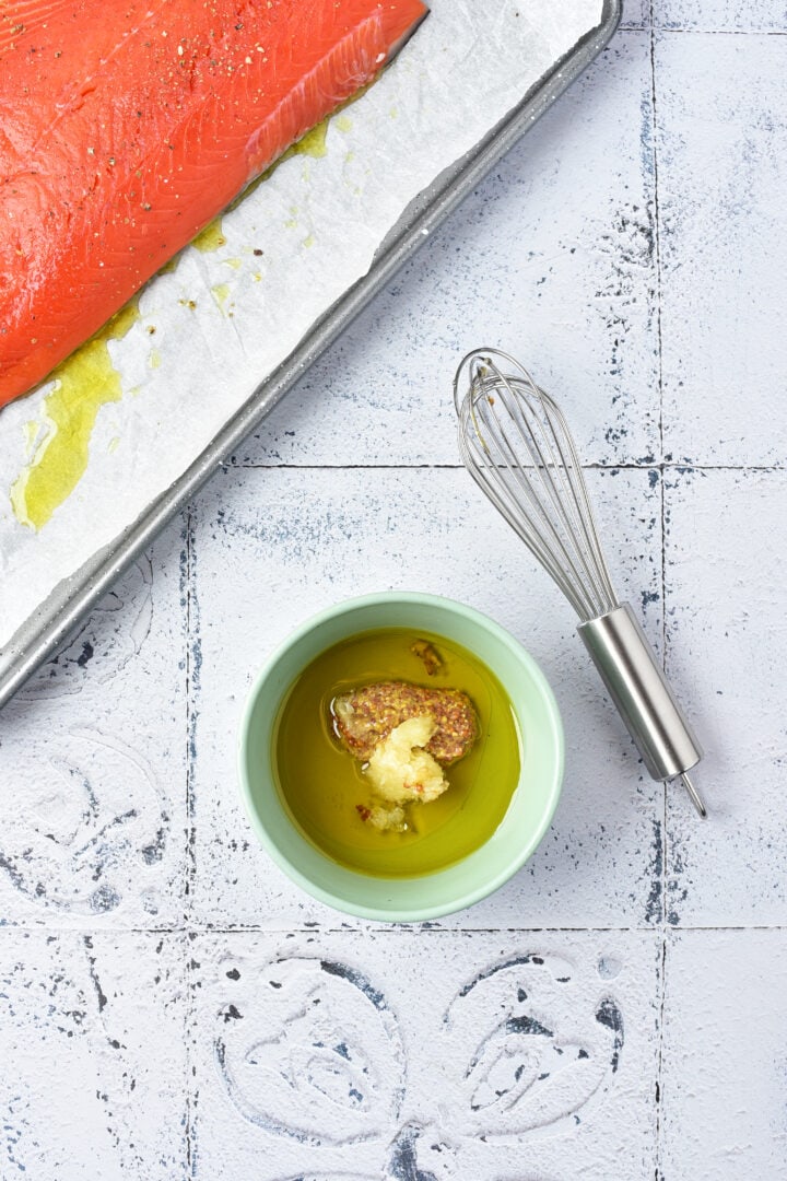 A bowl of oil with mustard and garlic, a whisk, and raw salmon on a baking tray with parchment paper.