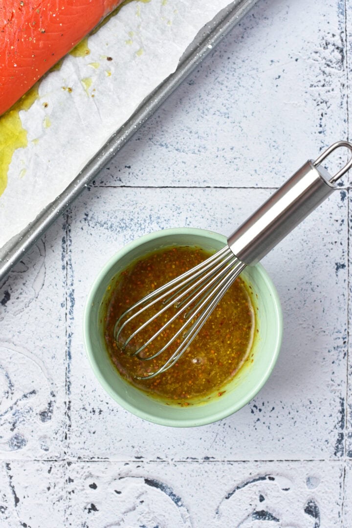 A bowl of mustard sauce with a whisk next to a tray of raw salmon on parchment paper.