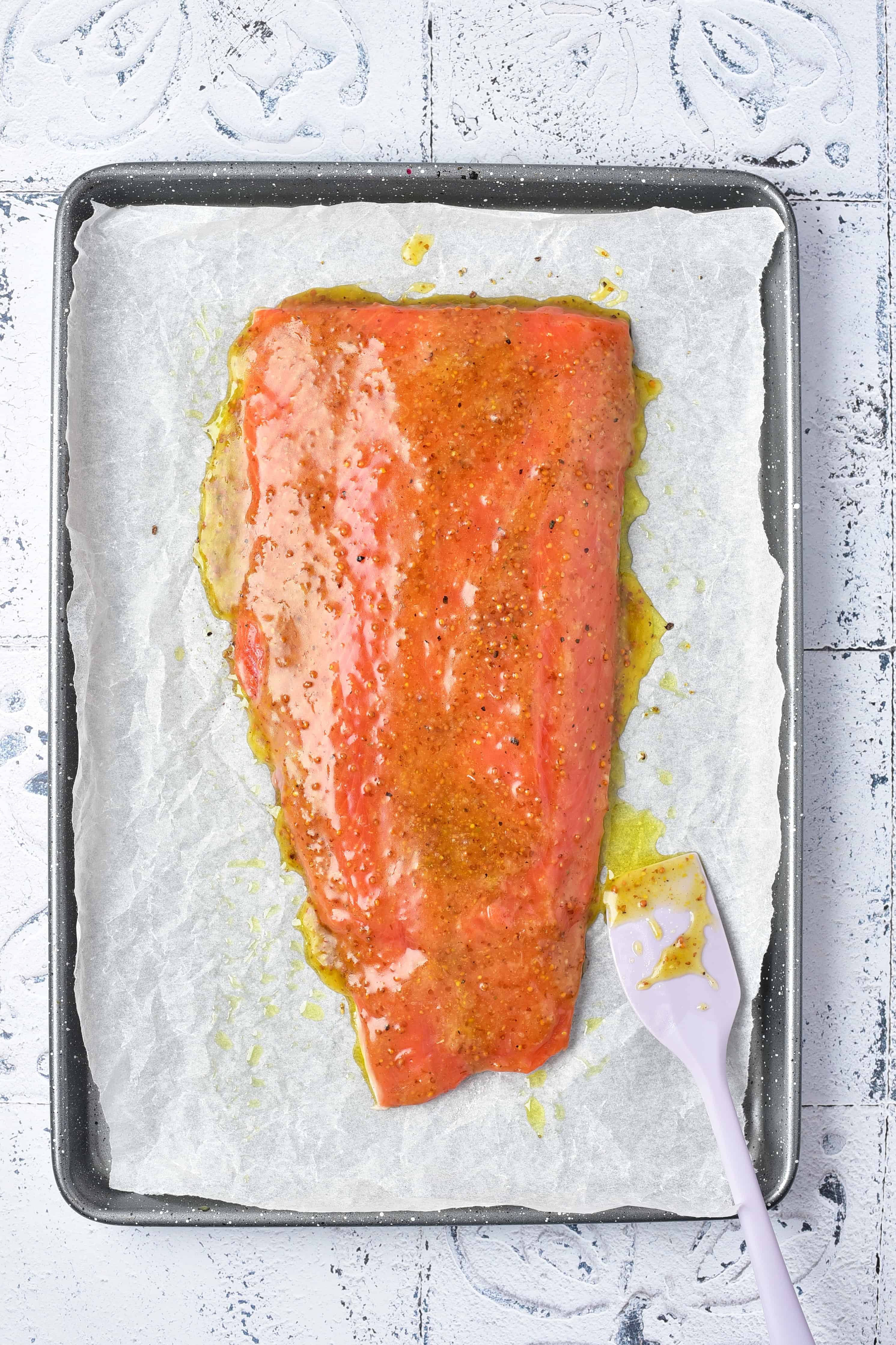 Raw salmon filet coated in marinade on parchment-lined baking tray with a spatula on the side.