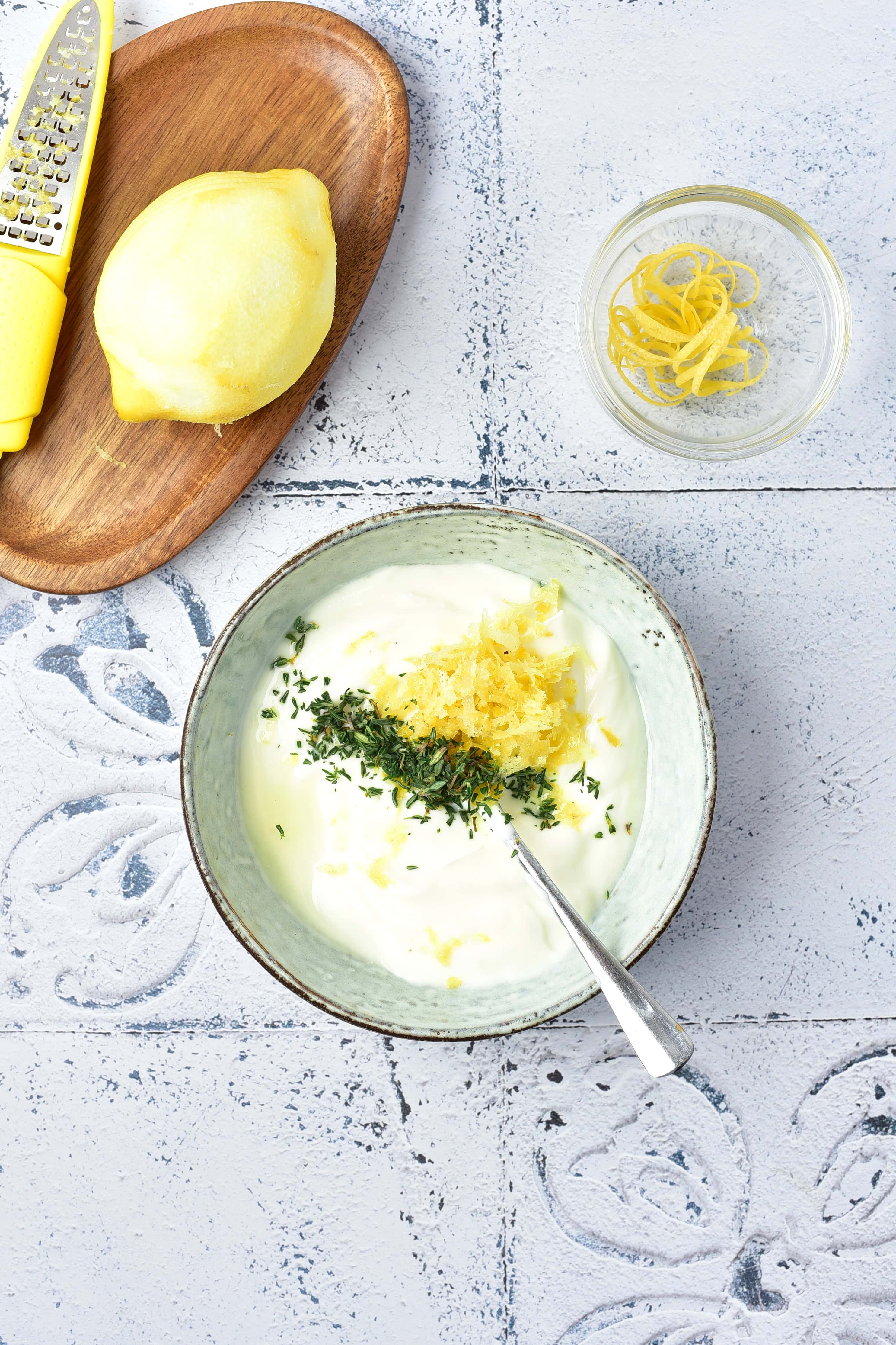 A bowl of yogurt with lemon zest and herbs beside a lemon, a grater, and a small bowl of lemon peel.