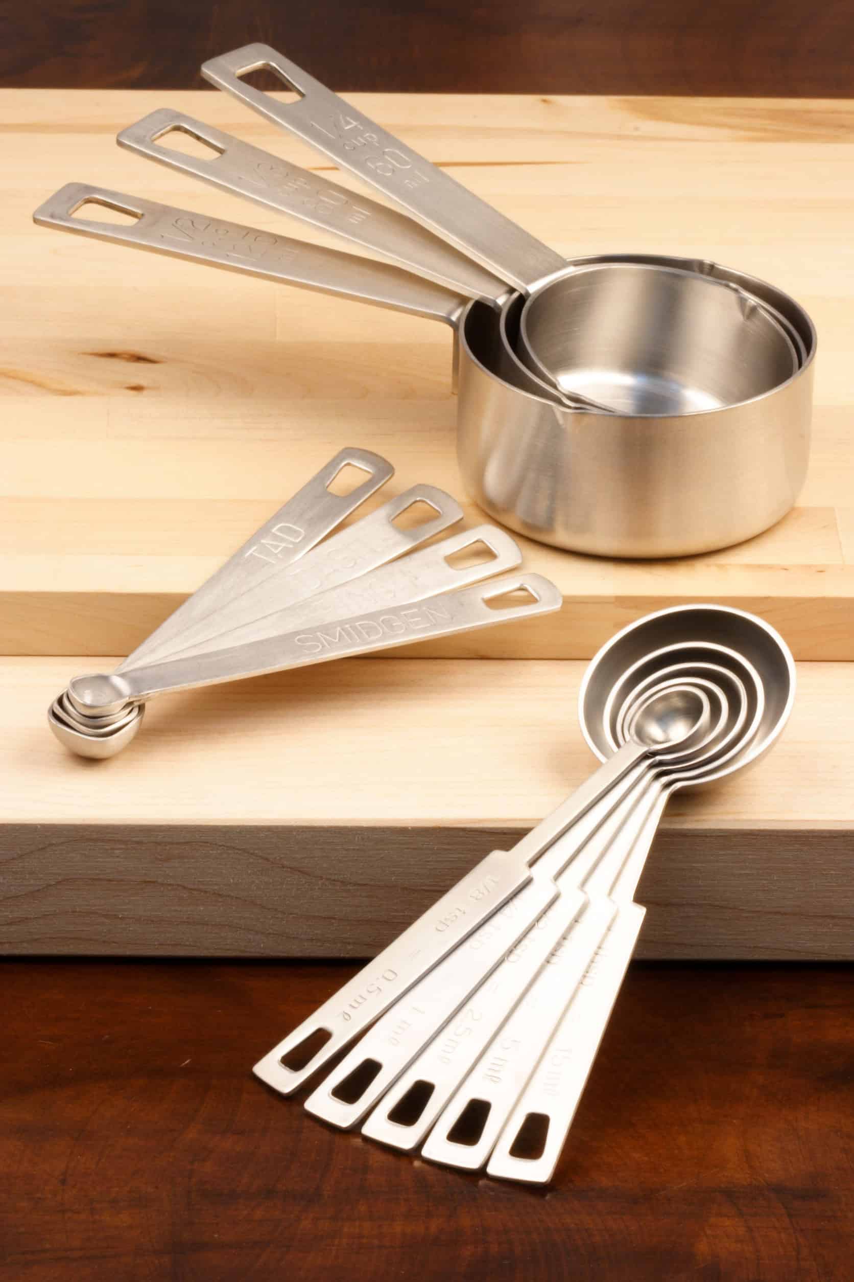 Metal measuring cups and spoons, perfect kitchen tools for holiday baking, rest on a wooden cutting board atop a dark wood surface.
