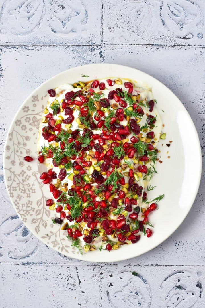 A plate of yogurt dip topped with pomegranate seeds, fresh herbs, and chopped pistachios on a patterned plate.
