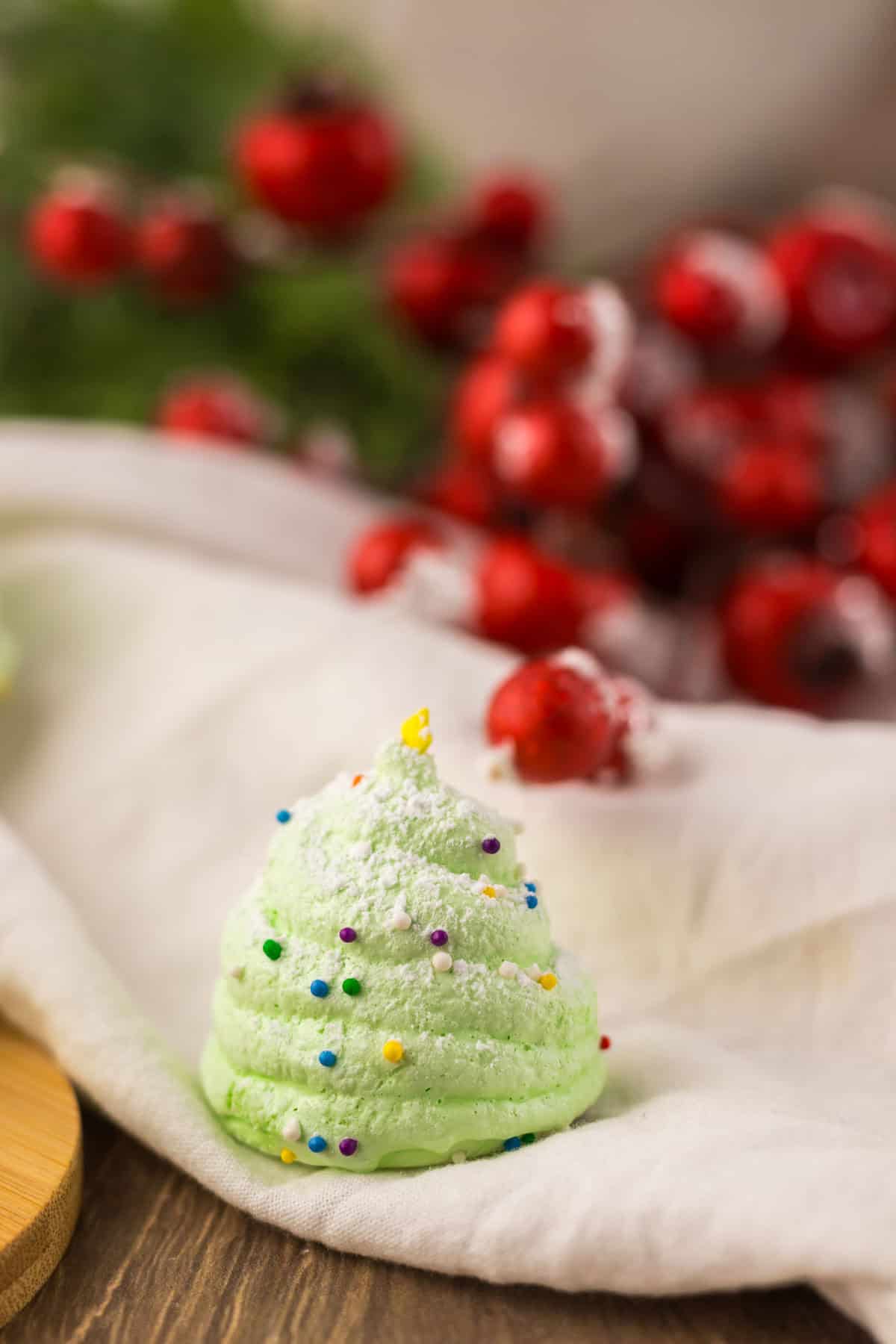 A green meringue shaped like a Christmas tree with sprinkles and powdered sugar, with red berries in the background.