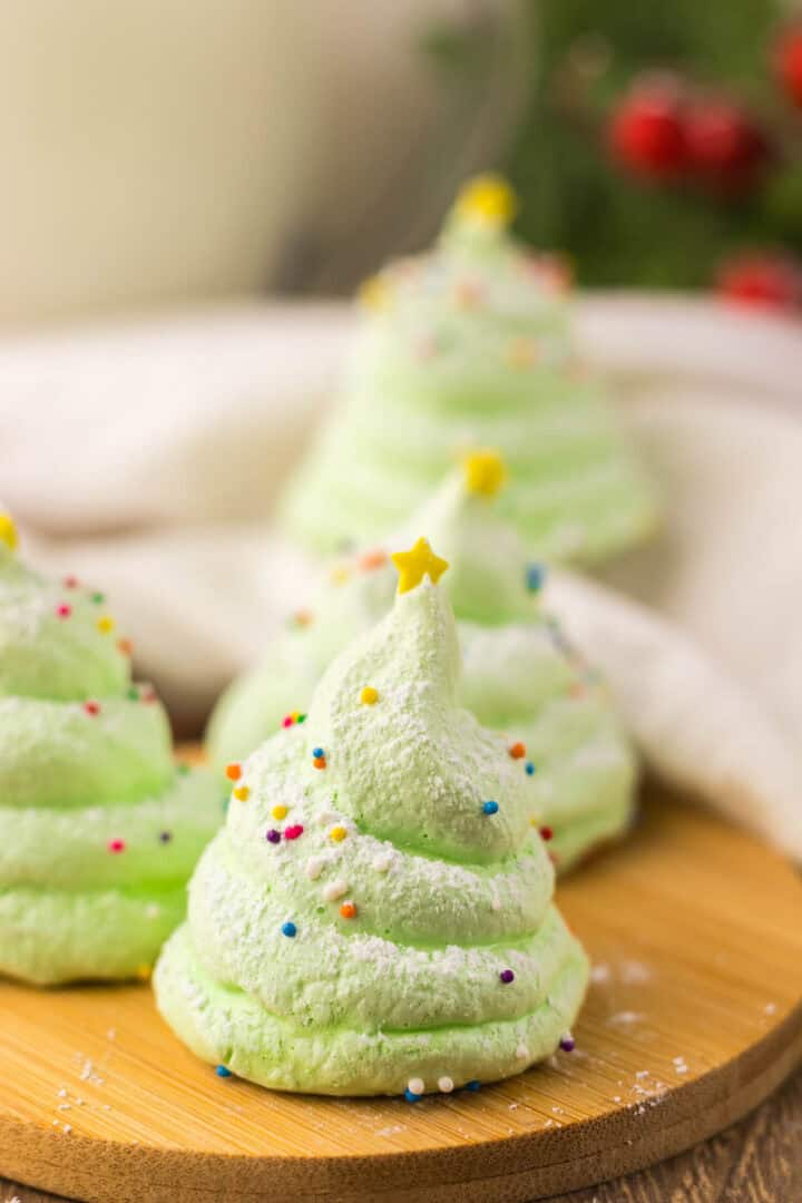 Green Christmas tree meringue cookies topped with powdered sugar, sprinkles, and a yellow star on a wooden board.